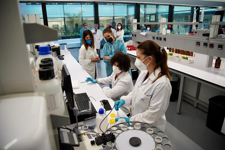 four women in masks and lab coats gathered around computer screen