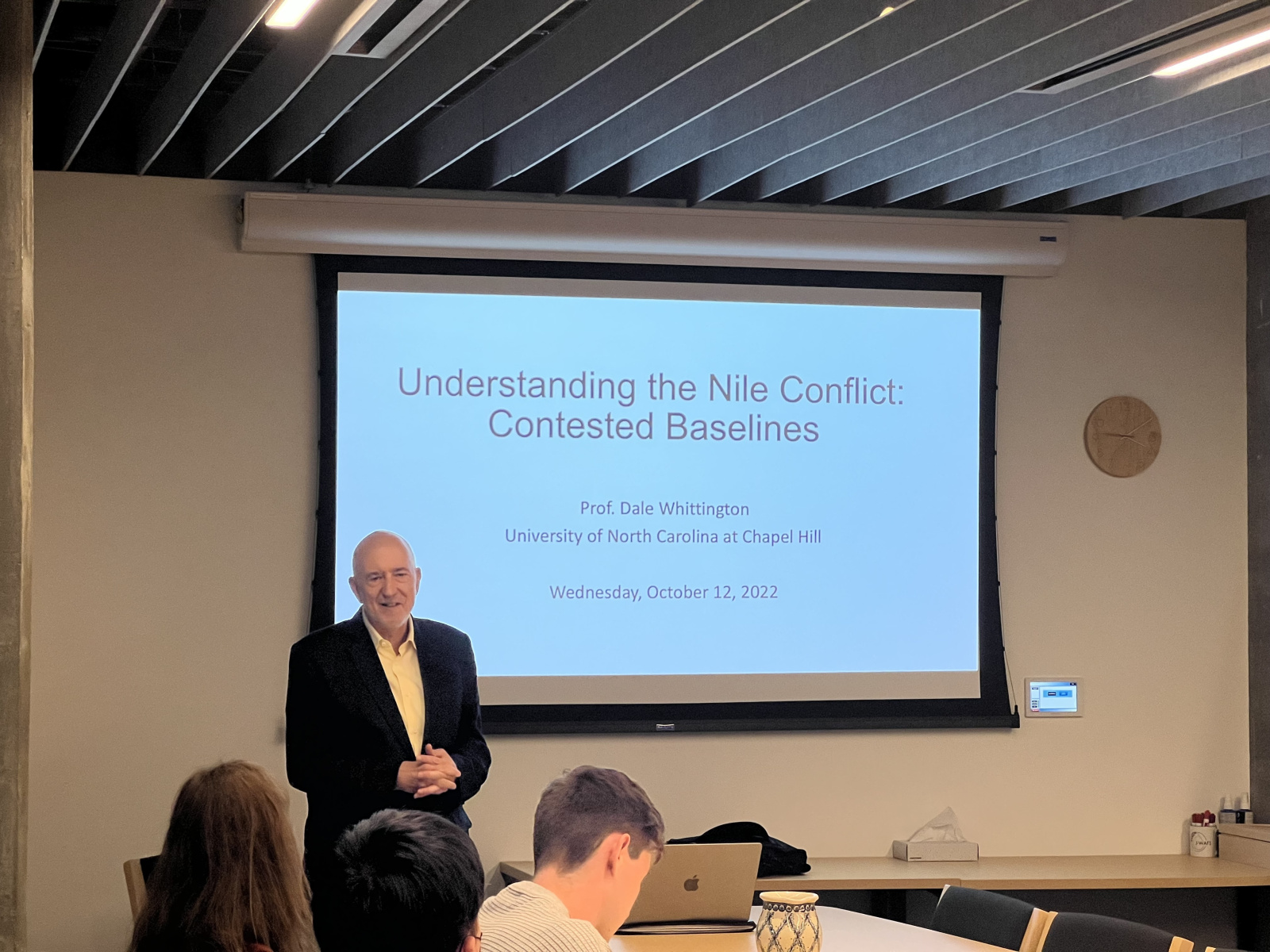 Dale Whittington giving a lecture with a presentation screen in back of him and people sitting at a conference table in front of him