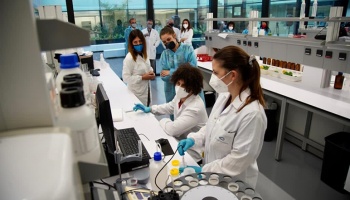 four women in masks and lab coats gathered around computer screen