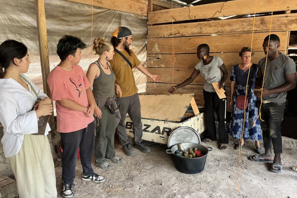 Seven people standing by a wooden box and a load of beeswax thermal batteries in a rough structure with a dirt floor