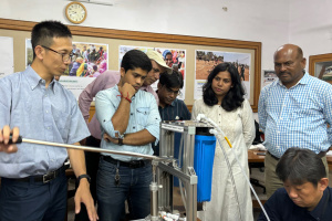 A group of seven people are gathered around the water-pump technology, watching a demonstration