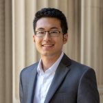 Asian man with dark hair and glasses wearing a suit in front of pillars