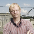 White man with red checkered shirt standing with arms folded outside in rural setting